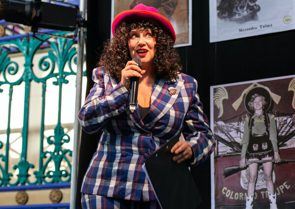 A person talking into a mocrophone, with brown curly hair, a red hat, and a blue and white checked suit