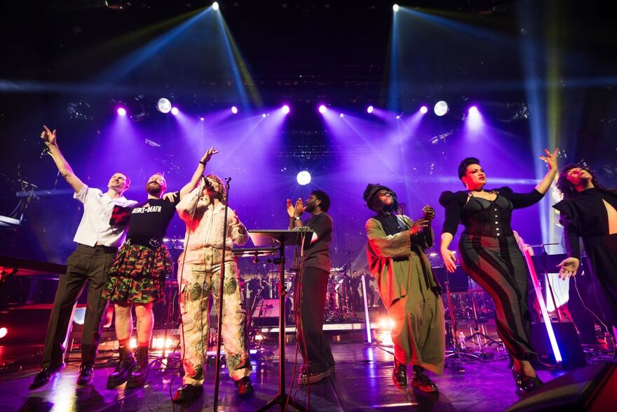 Performers stand on the front of the stage looking out at the audience, clapping and dancing