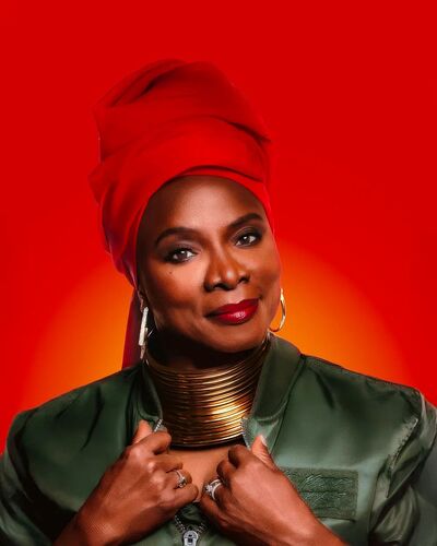 West African born Angélique Kidjo is visible from the shoulders up. She's wearing a red head scarf, gold multi-hoop necklace, gold hoop earrings and a green silk jacket. She's in front of a red and orange gradient backdrop