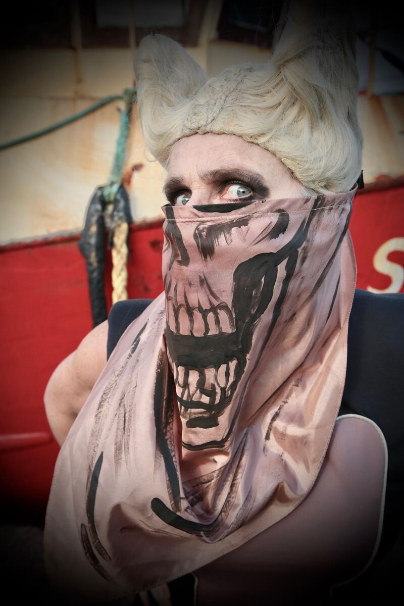 Liz Aggiss glares sideways with black eyeshadow, a blonde horned wig and a skull mouth scarf over her face
