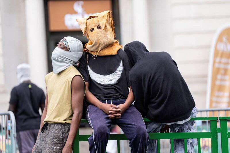 Three men sit or lean on a barrier, on with a grey head and face covering, one wearing a hessian bag with eyeholes cut out, and one wearing a black hoodie with his back to the camera