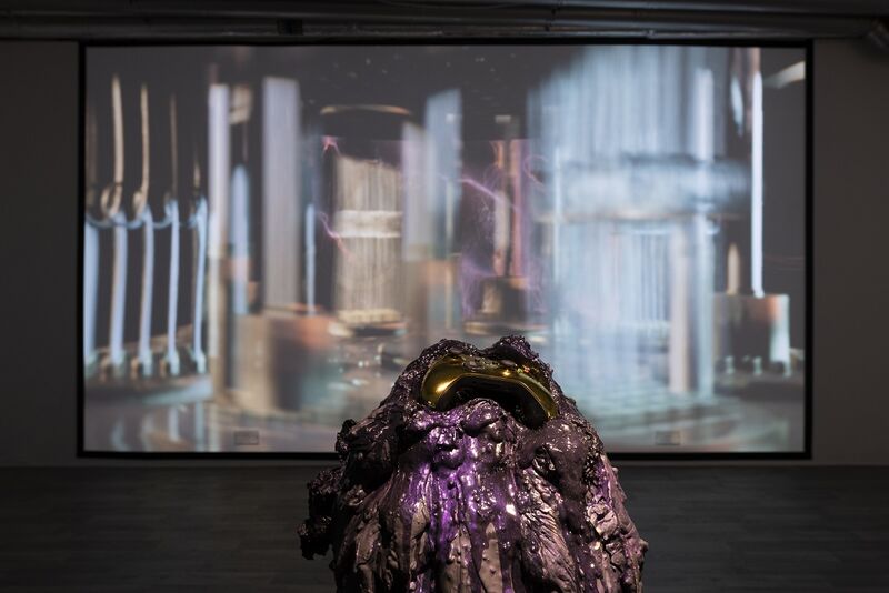 A close up of a purple, oozing sculpture with a gold video game controller pressed in to it. Behind the sculpture is a large digital screen