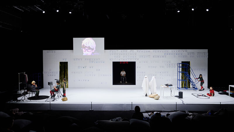 A wide shot of the white staging shows various children on stage, including two dressed as ghosts in the centre. There is a man sat in darkness in the wall at the back of the stage