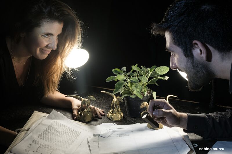 Two actors stamp paperwork on a tabletop, pupeteering plastic dinosaurs