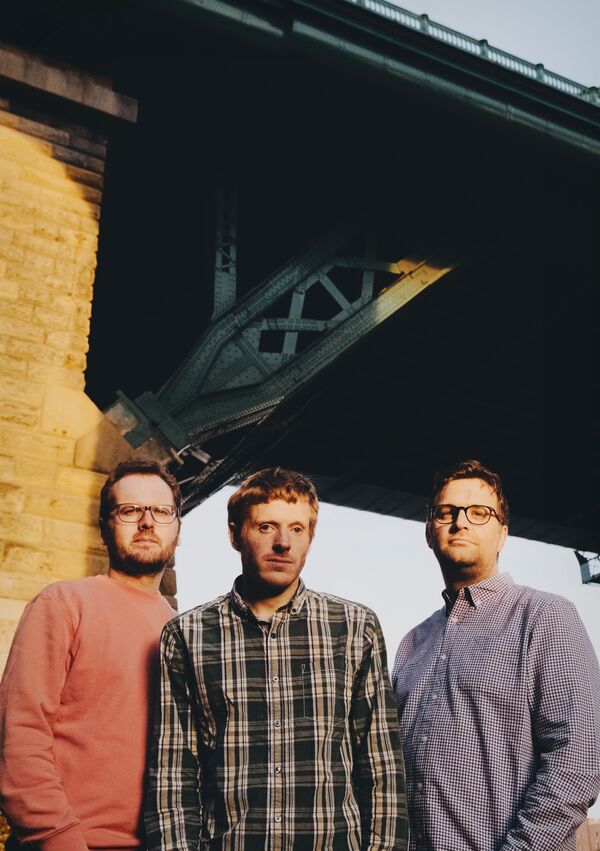 Three men stand under a bridge looking directly at the camera. They all have short dark hair, and two of them wear glasses.