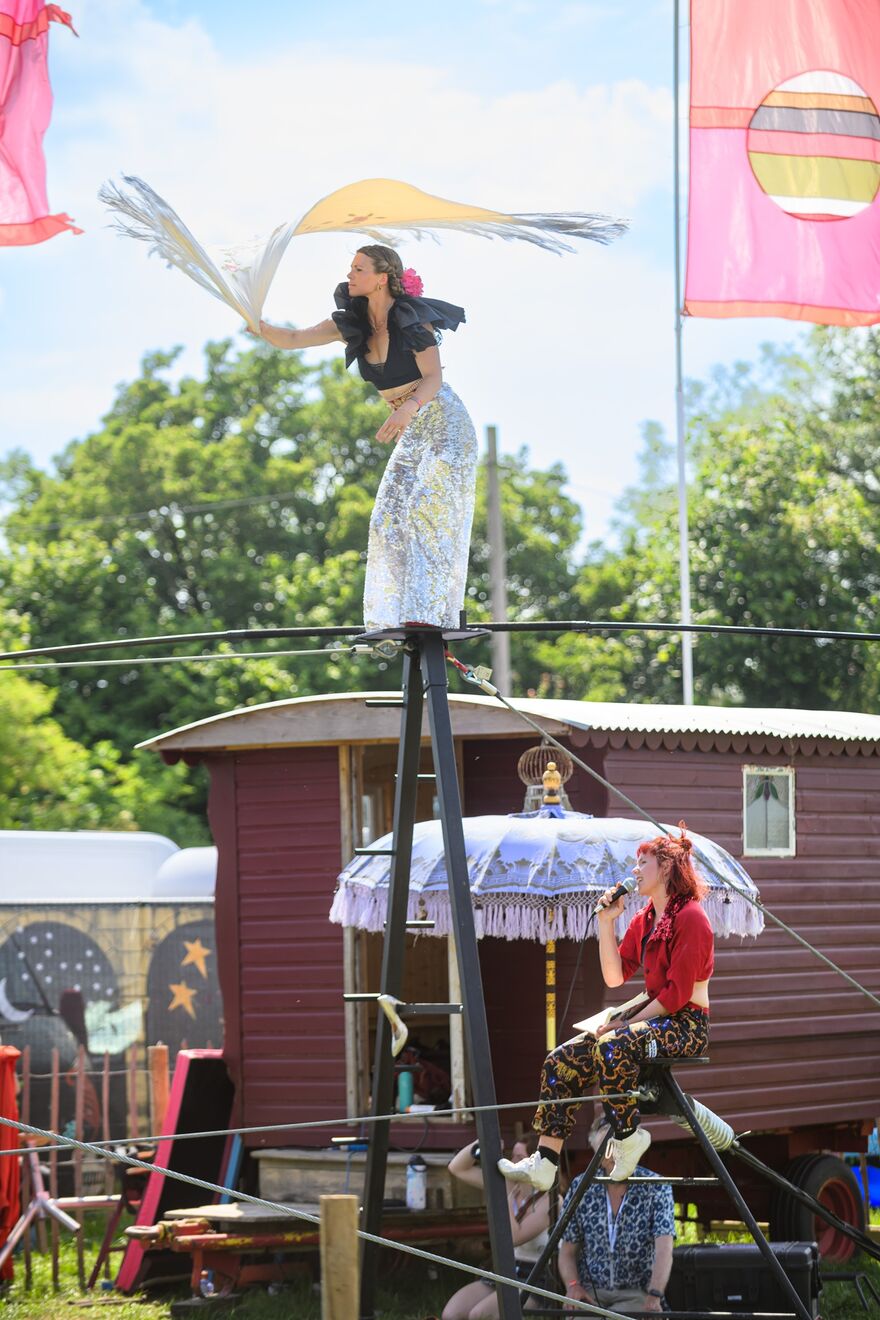 A woman in sparkly silver trousers and a black top stands on a high wire whilst another woman sitting on a high stool speaks in to a microphone, they appear to be at a festival, with flags and a caravan in the background