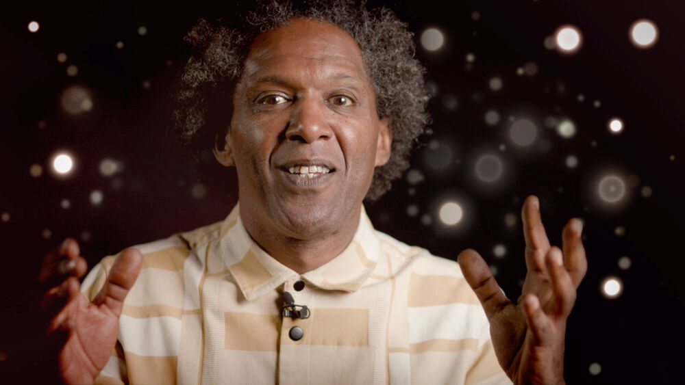 Lemn Sissay sits infront of a dark background, lit up with many sparkling star-like dots  