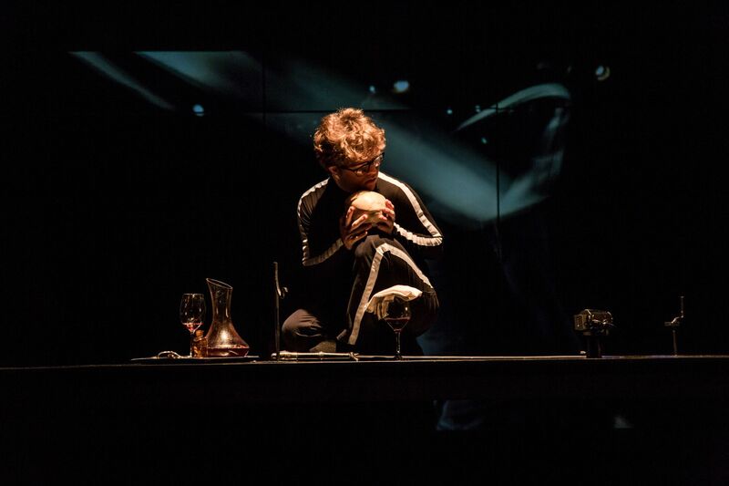 An actor crouches on stage, drak and shadowy lighting, he holds a skull with glasses and a carafe of wine around him