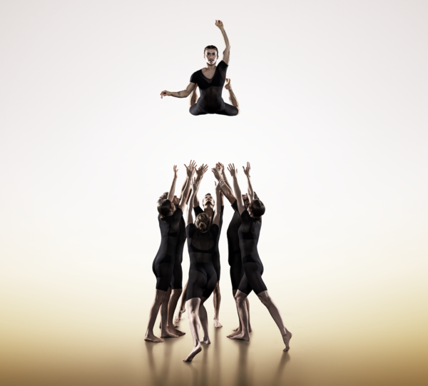 A group of acrobats in black unitards have thrown another acrobat high in the air, the are standing below him, with their arms raised, ready to catch him