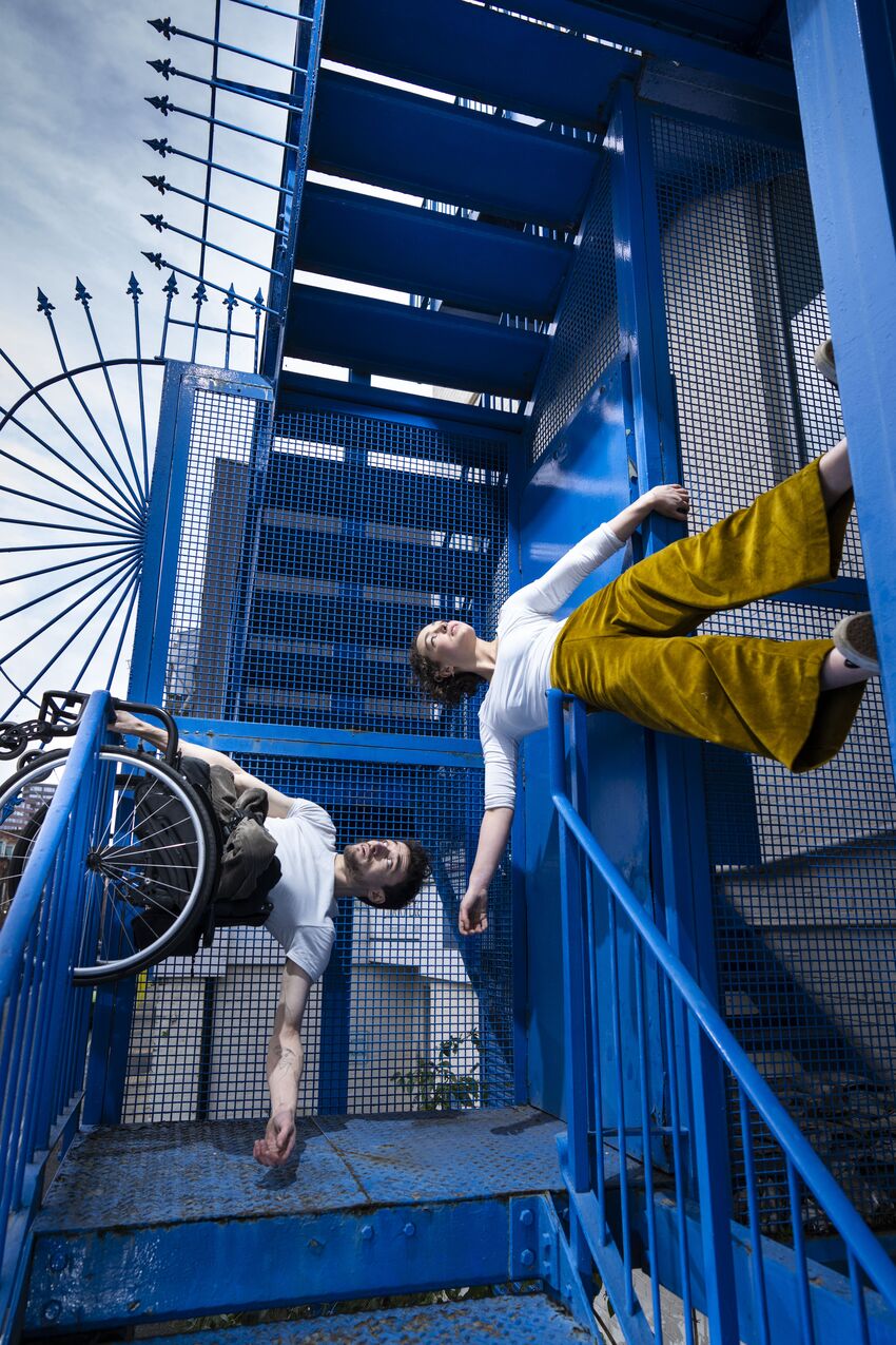 On a blue staircase, a man in a wheelchair is hanging off the railings and a girl is resting her waist on a bannister
