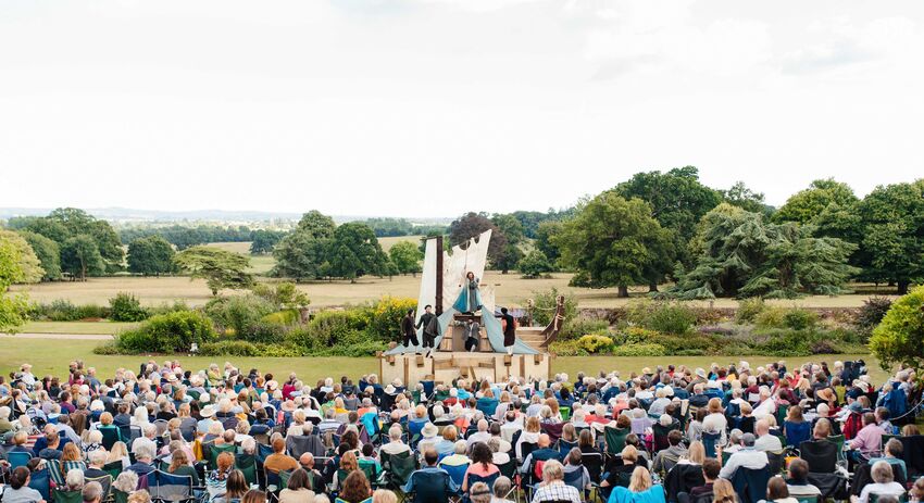 A large group of people sitting on camping chairs are watching a performance against a backdrop of trees and fields