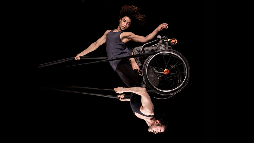 Black background. A male and female performer swinging in the air like a pendulum, reaching it's highest point, where the male performer in a wheelchair hangs upside-down, with the female performer upright leaning on him, both attached to black circus straps