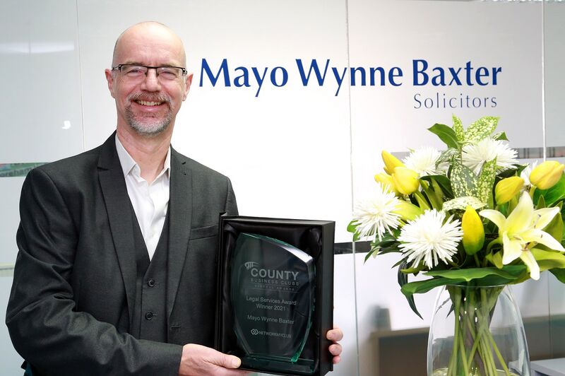 A bald, white man in glasses holding up an award next to a bunch of yellow and white flowers