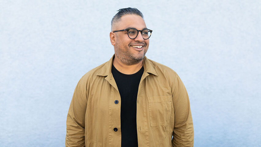 Photo of Nikesh Shukla smiling, standing in front of a white wall wearing round glasses a black top and camel colour jacket