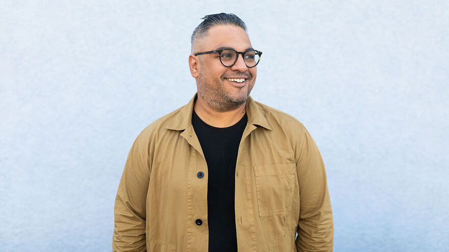 Photo of Nikesh Shukla smiling, standing in front of a white wall wearing round glasses a black top and camel colour jacket