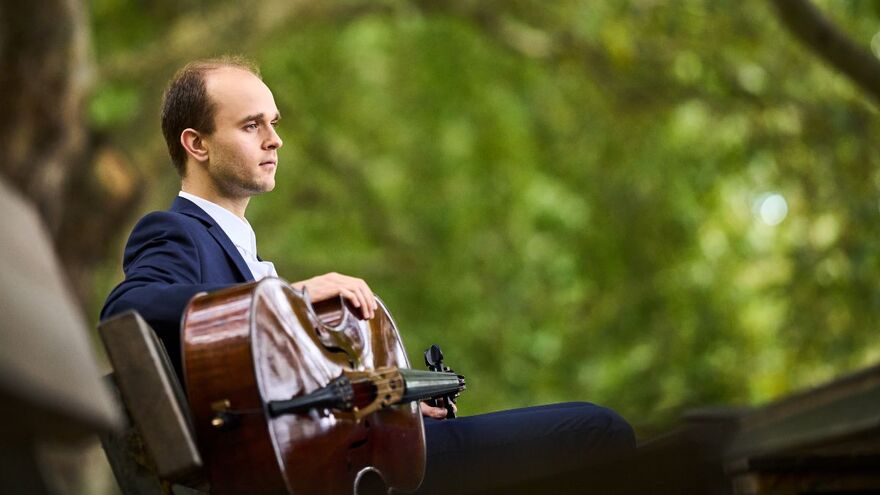 Maciej Kułakowski sits in a forrest. He has his cello over his lap and stares into the middle distance
