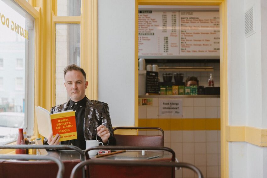 Inside a cafe, a man wearing a black sequin jacket holds a book called 'Inner Secrets of Card Magic' while stirring a coffee cup 