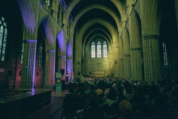 People sitting watching a musician on stage. They are inside a church with large concrete poles running throughout and a big triple window at the end of the room