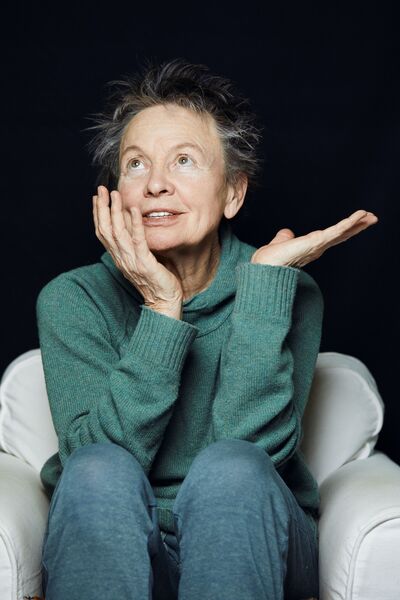 Laurie Anderson- a 78 year old woman with short grey hair is wearing a teal knitted jumper and blue trousers. She's sat in an armchair with her head in her hand and staring towards the ceiling