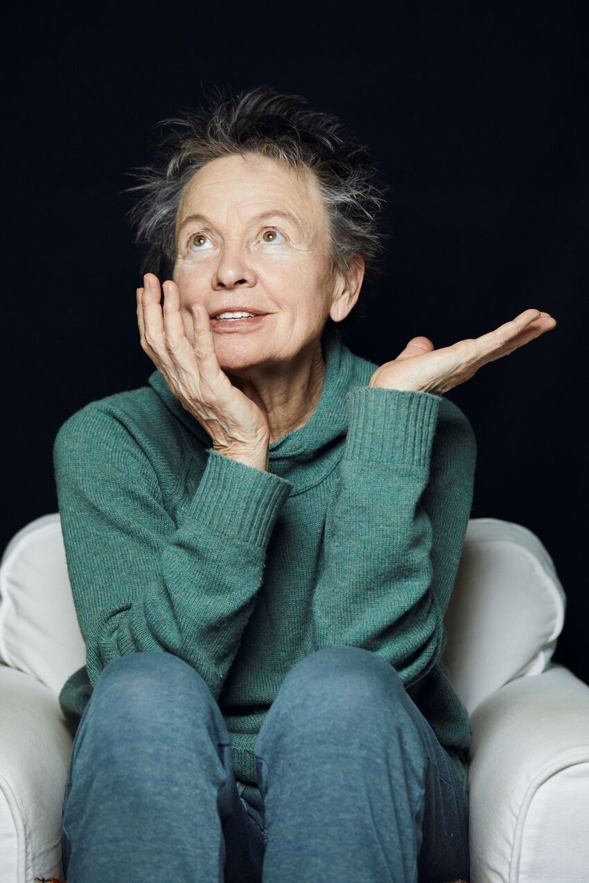 Laurie Anderson- a 78 year old woman with short grey hair is wearing a teal knitted jumper and blue trousers. She's sat in an armchair with her head in her hand and staring towards the ceiling