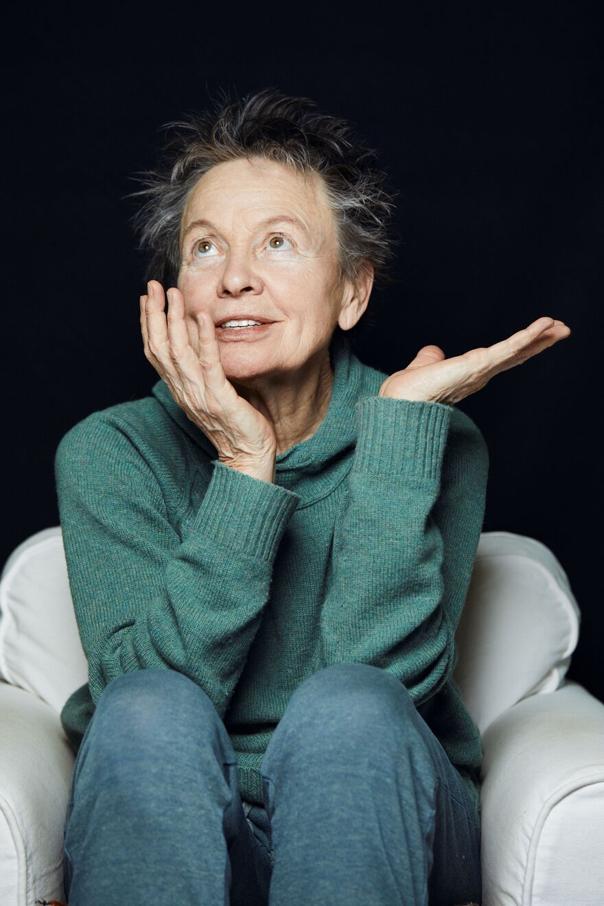 Laurie Anderson- a 78 year old woman with short grey hair is wearing a teal knitted jumper and blue trousers. She's sat in an armchair with her head in her hand and staring towards the ceiling