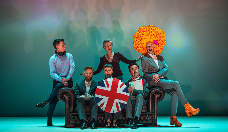 The cast of Abomination: A DUP Opera sit on a coach on stage against a hazy turquoise background. One cast member holds a shield with the Union Flag on it. Another wears a giant wig that resembles a marigold.