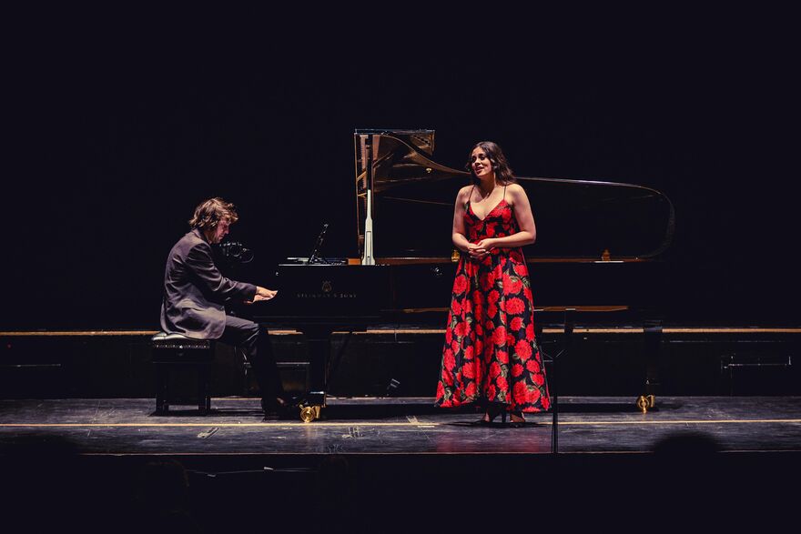 Jerwood Youngs Artists event from 2021 - a man in a black suit plays piano on stage, with a woman waring a long black dress with red flowers sings at the front of the stage