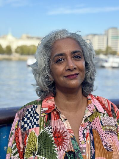 An Indian woman with mid-length grey hair, wearing a brightly coloured shirt stands in front of a body of water