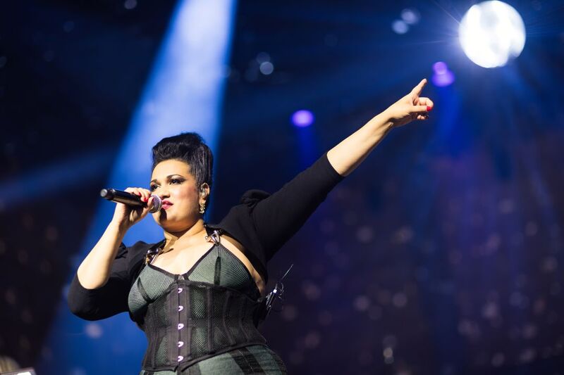 On-stage portrait of a performer singing into a mic and pointing with their left arm