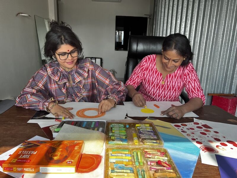Two women sit at a table drawing circles on white paper. One is using orange, and the other, yellow.