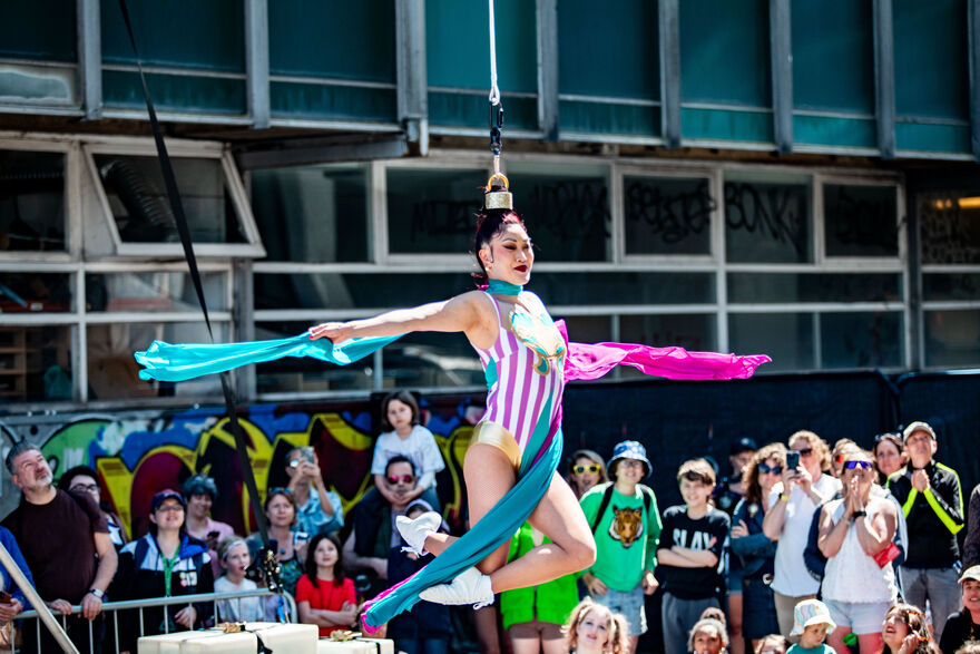 A showwoman in pink and blue striped leotard hangs from her hair, her arms outstreched as an audience watches on