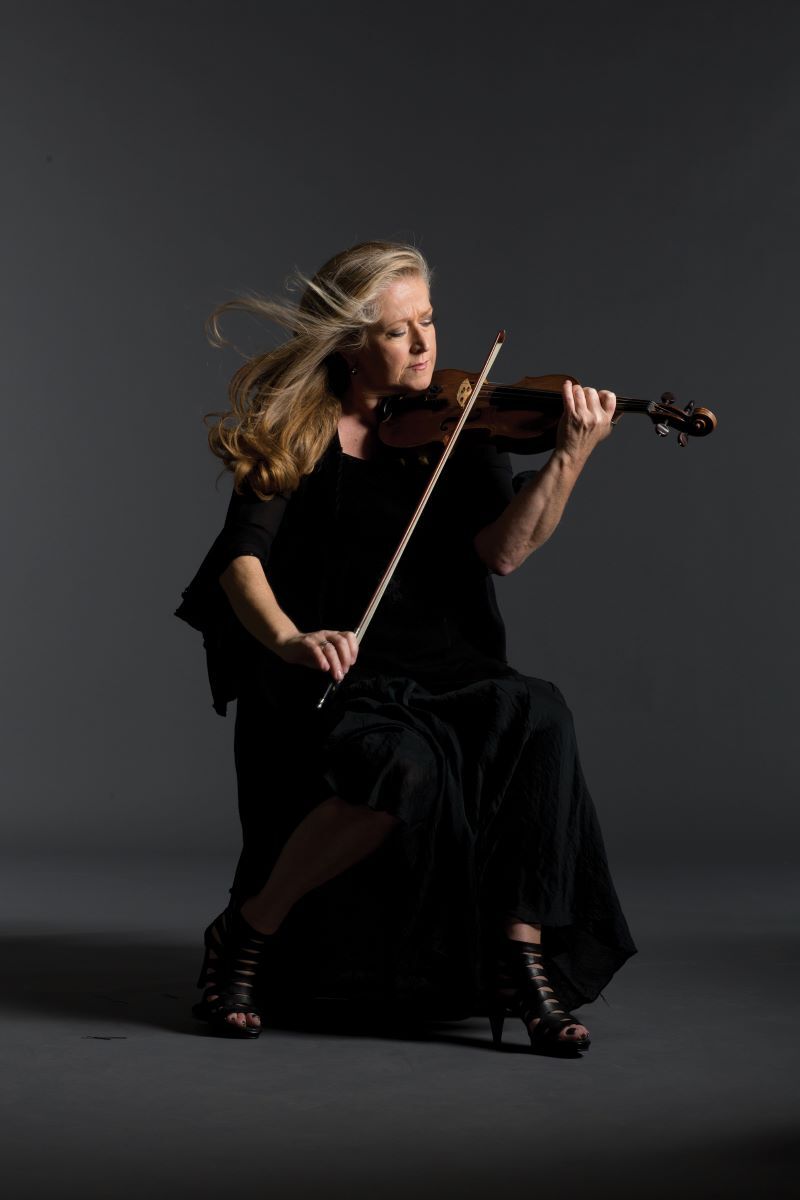 Jacqueline Shave sitting playing the violin, wearing a long black dress with wind blowing her hair back