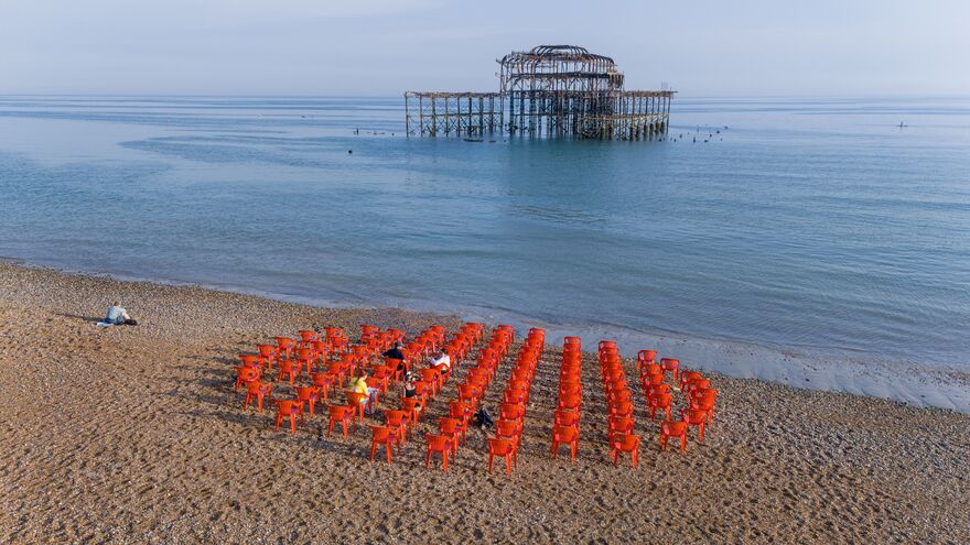 A circle of red chairs on a pebbly beach in front of the west pier