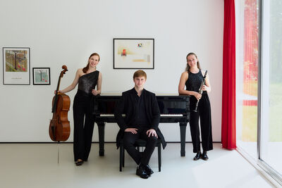 Delphine Trio in a bright room with a grand piano. Someone is sitting on the piano stall and two musicians are standing either side, with on holding a cello and another holding a clarinet