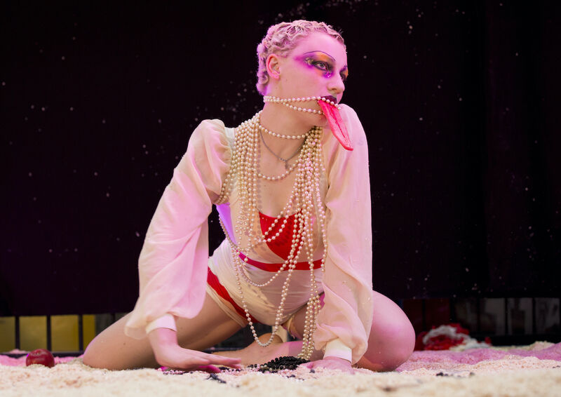 A person with pale skin wearing a light coloured dress and lots of pearl necklaces crouches on the ground, with red eye make-up and a large fake tongue hanging from their mouth