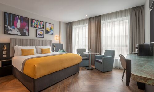 A hotel room with a large king size bed, nice artwork on the walls, a table with two chairs and lovely large windows