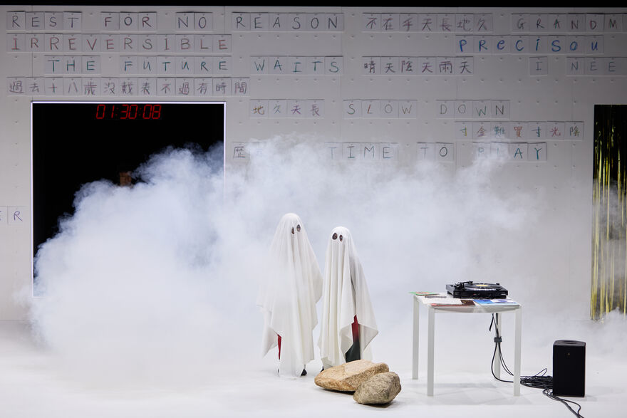 Two children dressed as ghosts in a cloud of smoke in front of a white wall covered with sheets of paper with handwritten letters