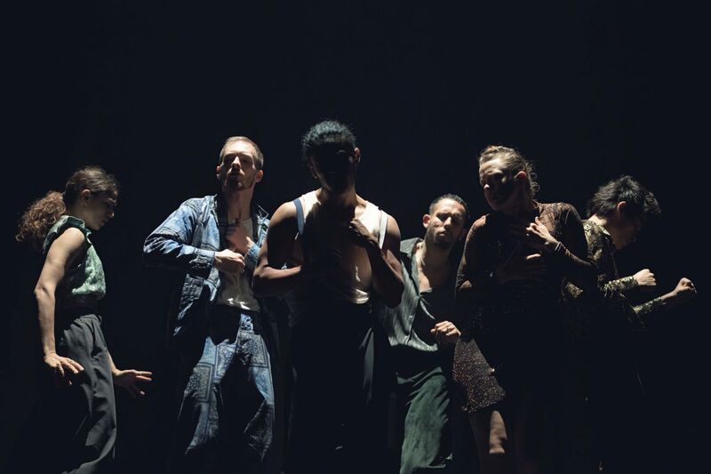 11.	6 dancers move in close proximity to each other, with 2 dancers in the foreground and 4 dancers slightly behind them. They are all performing slightly different moves, with some raising their arms and leaning forwards, and another stretching her arms towards the flooring in a thrusting motion. Their facial expressions are content, and they look as if they are dancing in their own world – they are not interacting with one another. They are lit by a single white light, and are dancing in and out of shadows. They are dressed in an assortment of colourful shirts, trousers and dresses.