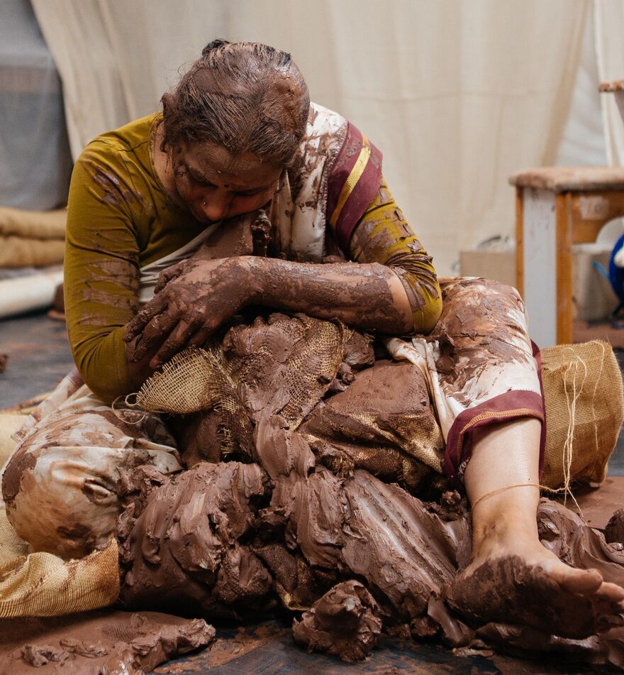 Performer Vidya Thirunarayan sits cross legged on the floor wearing traditional Indian dress and covered in clay