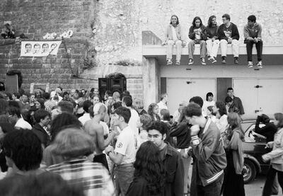 A group of ravers are partying below a cliff edge, there is a building to the right, with a group of 5 people sitting on the roof