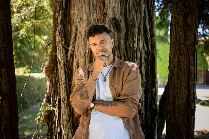 A picture of Raymond Antrobus standing in front of a tree, his hand on his chin