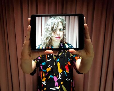 A person in a colourful shirt holds a tablet in front of their face, you can see a picture of their face on the screen of the tablet