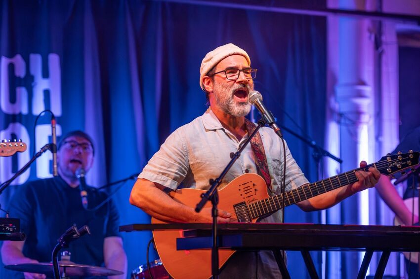 Adam Buxton playing a guitar and singing on stage