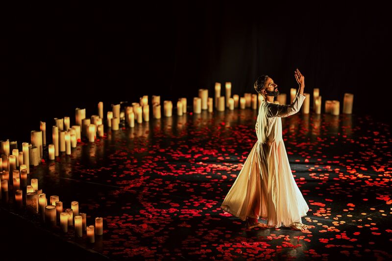 A man in a white dress stands before a semi circle of large white candles. He holds his right hand up. Beneath him is a black floor covered in red petals.