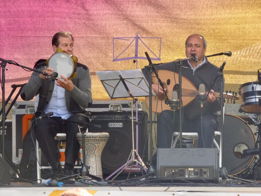 Two people are on stage, seated. One is playing a tambourine and the other a lute.