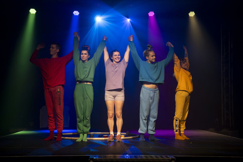 Five performers stand on stage. They all wear costumes of one block colour, and the lighting overhead mirrors this
