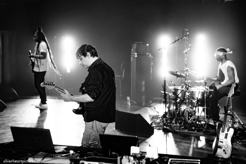 Black and white photo of 3 musicians on stage standing sideways on. Left to right there's a Black man with long braided hair playing guitar, a white man with short dark hair playing bass, and a Black man wearing a bandana on drums