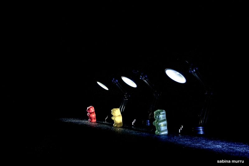 Three Gummy Bears in a row, red on the left, yellow centre and green right, each spotlighted by a small torch in a very dark setting