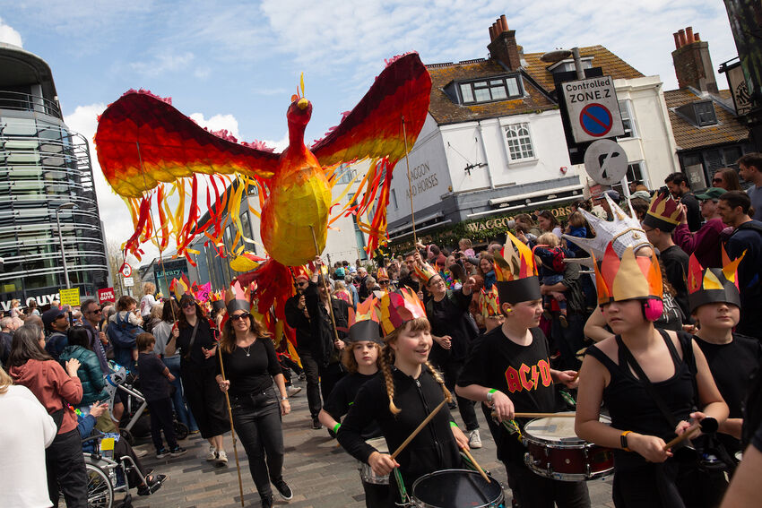 A group of primary school children are parading through the streets of Brighton. They are banging drums and a giant phoenix sculpture flys above them