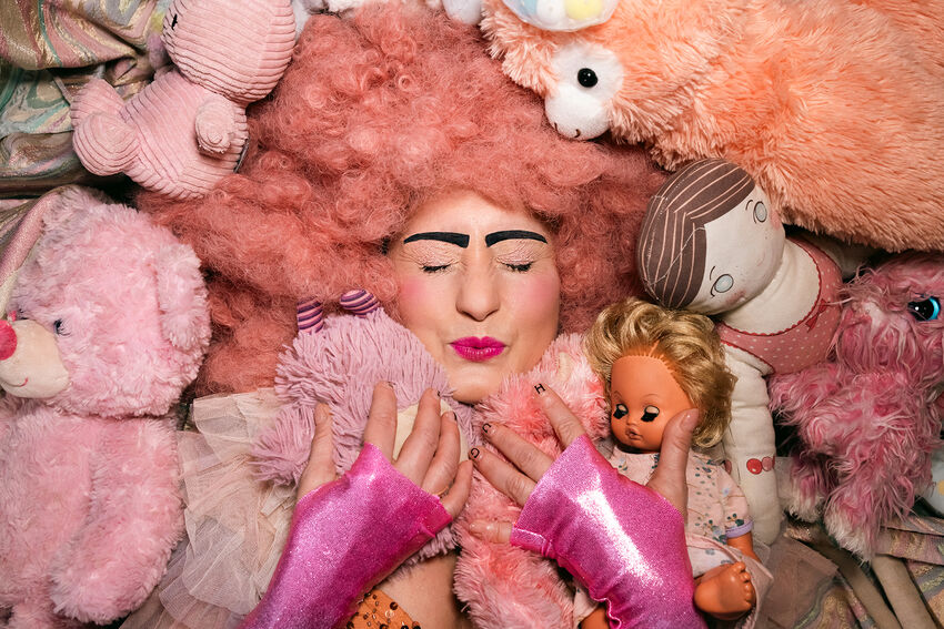 A performer wears a bright pink wig and a pink dress. They are enveloped in a hug of many arms, although you cannot see who is hugging them